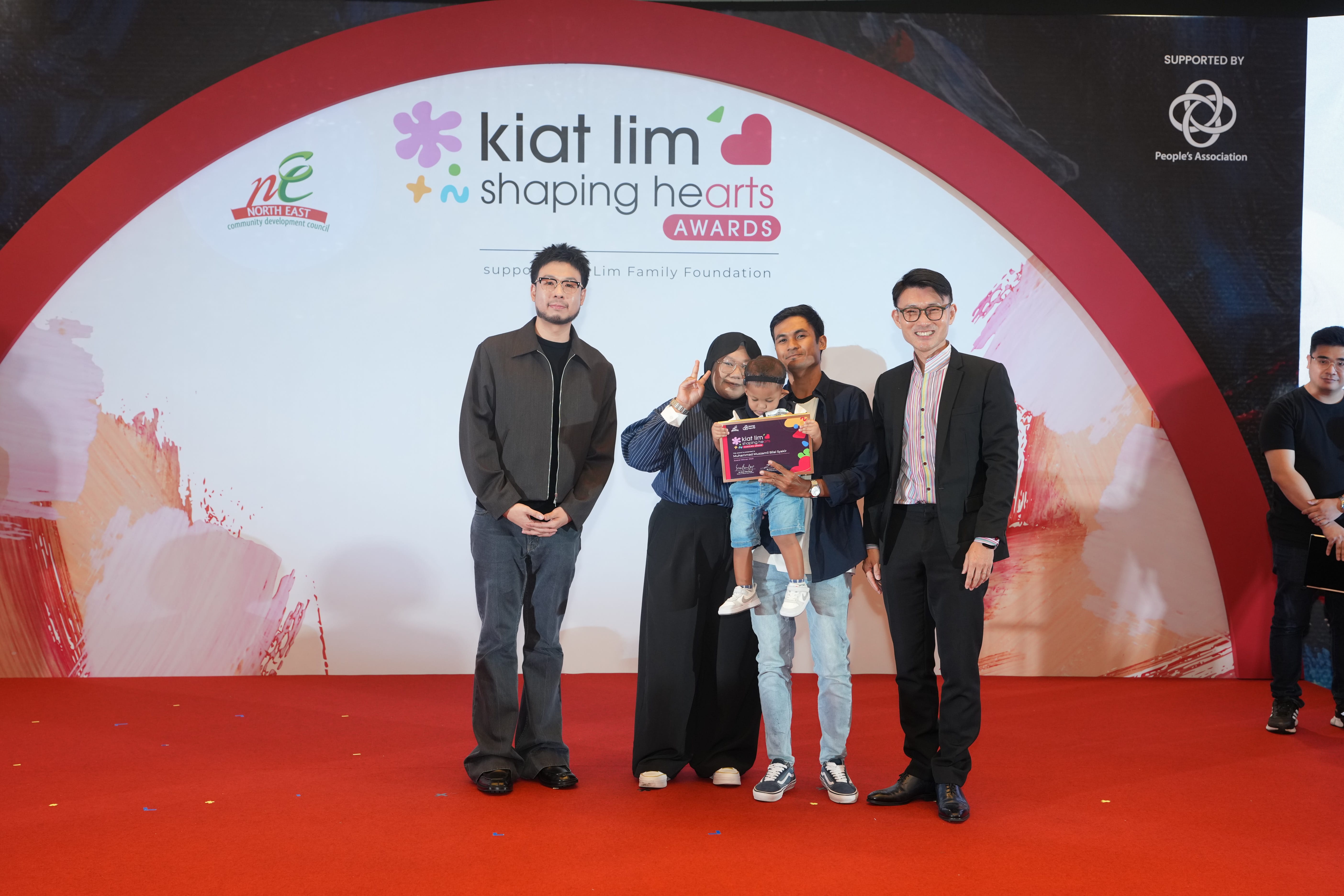 Award recipient standing on stage with the presenter, holding their certificate and posing for a group photo during the award ceremony, with a backdrop reading ‘Kiat Lim-Shaping Hearts Awards'.